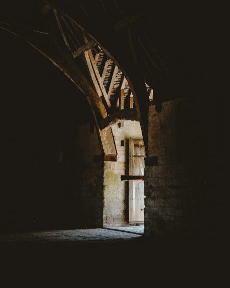 Dark stone archway with wooden beams leading to a brighter interior.